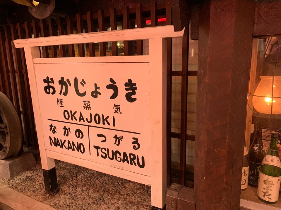木製の看板に「おかじょうき 陸蒸気 OKAJOKI なかの NAKANO つがる TSUGARU」と書かれた駅名標風サイン、奥に日本酒瓶