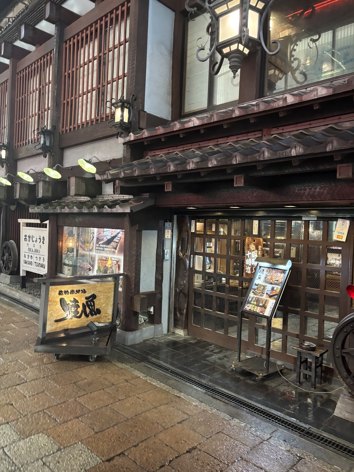濡れた石畳の前に立つ陸蒸気の入口。格子戸と鉄製ランタン、木製の看板と店内写真のメニューボード