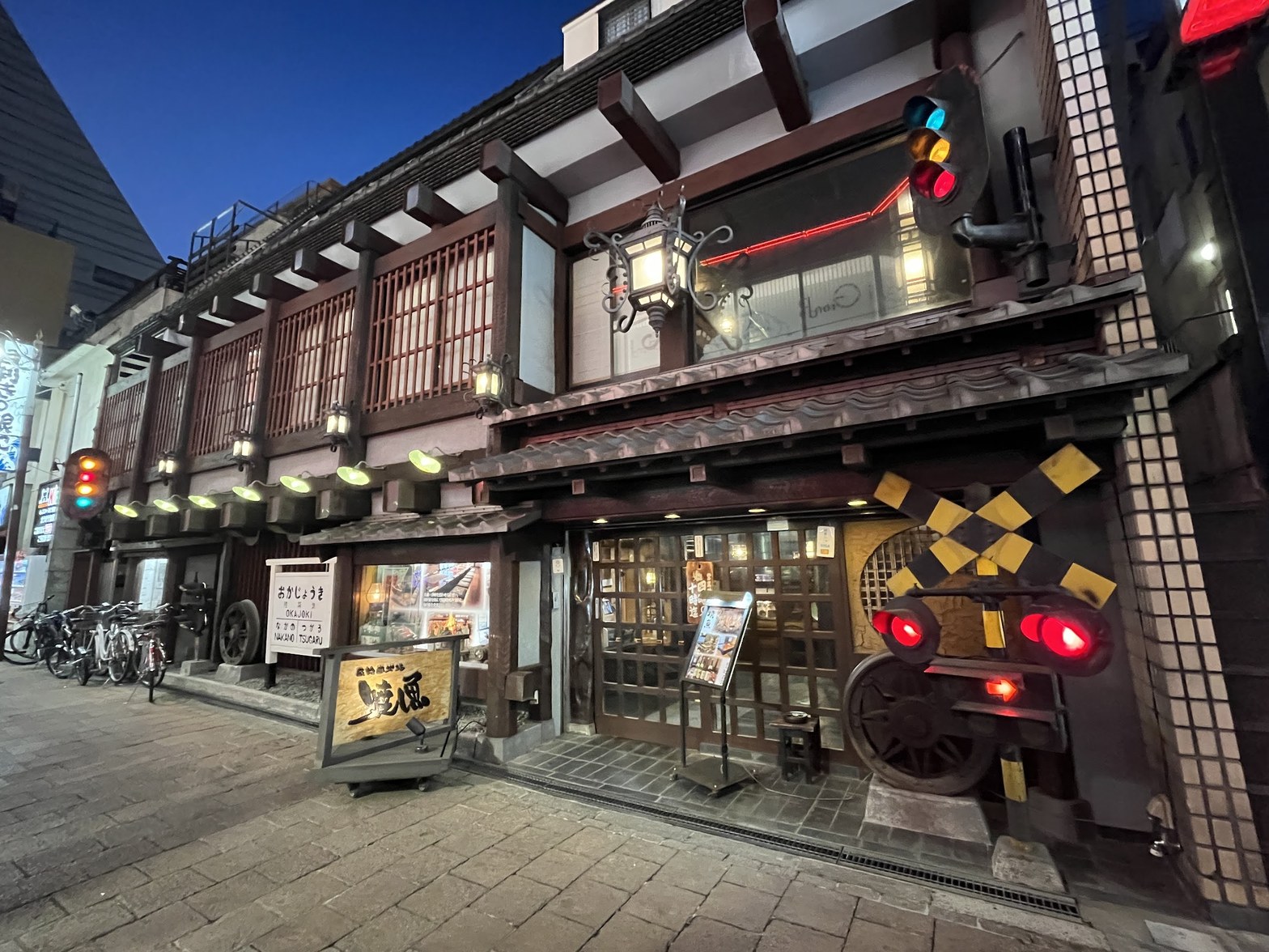 夜の街灯に照らされた陸蒸気の外観全景。瓦屋根の格子窓、蒸気機関車の車輪を模したオブジェ、赤と青のランプが並ぶ